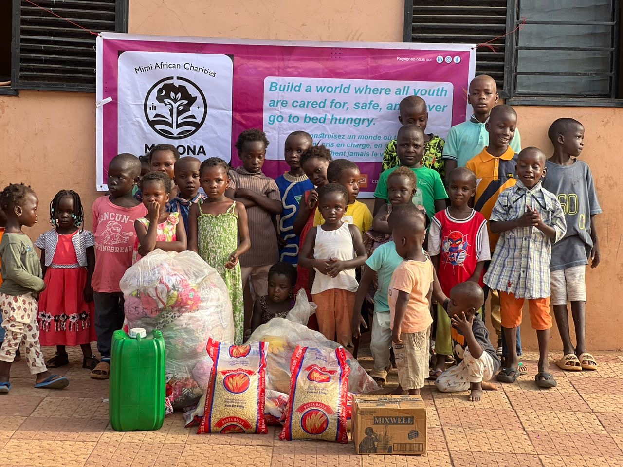 MACONA distribution event in Africa: about 25 children grouped in front of the MACONA banner with donated rice, food, and supplies in the foreground
